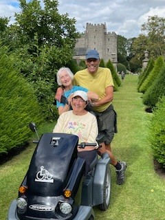 An image of Jean sat on a Tramper, facing the camera. It is a summer's day and she is wearing a cap and t-shirt. A woman and man are standing behind Jean, smiling to the camera. There is greenery to each side of them and an imposing grey structure in the background.