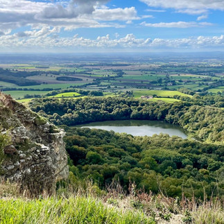 A beautiful vista, taken by Jilly. It depicts lush green countryside, with a body of water central. The sky is blue and cloudy.