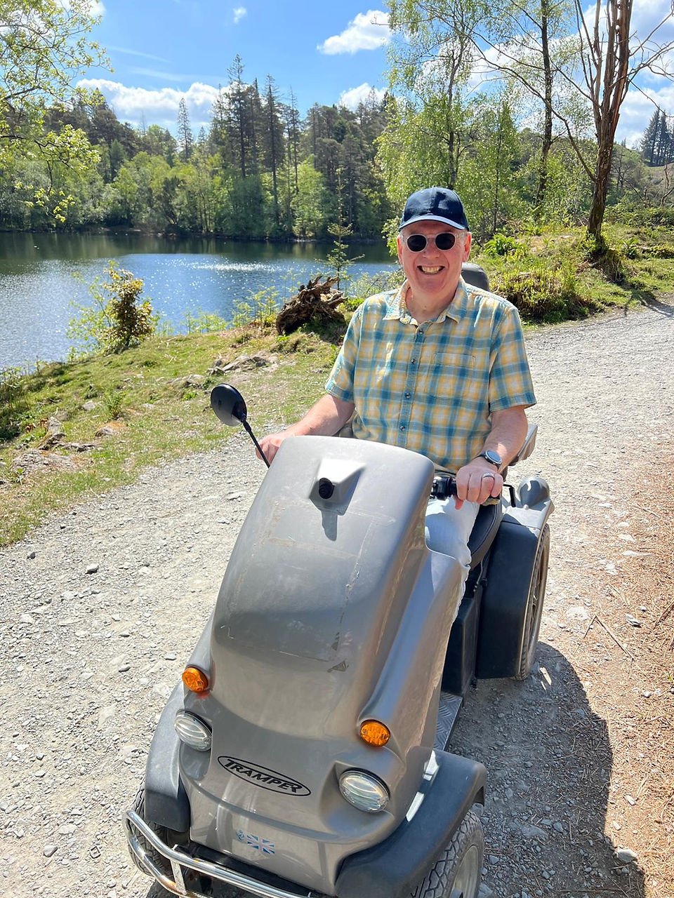 An image of Chris on a silver Tramper. He is smiling to the camera and wears a short sleeve, checked shirt, a baseball cap and sunglasses. Behind him is a body of water, with grass verges and woodland in the distance. 