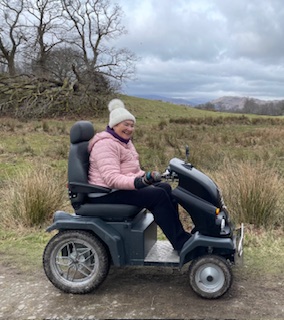  A side view image of Jean on a Tramper. It is an autumnal day with barren grass and bare trees in the distance. Jean wears a pink coat and a cream bobble hat.