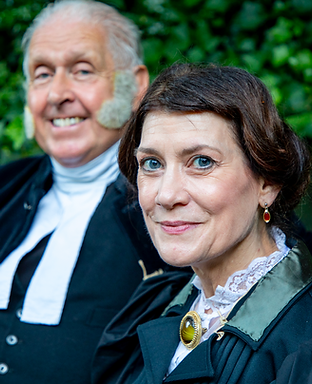Two performers are dressed in Victorian clothing. One, an elderly man and the other a lady, smile into the camera warmly.
