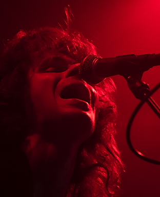 Joel of 'The Shucks' performs at the Norwich Arts Centre. He is singing into the microphone and is lit in red.
