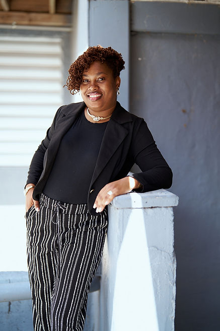 Sandy Lewis, dressed in black stripped pants and black shirt with a black business jacket leaning on a cement ledge looking at the camera with a smile