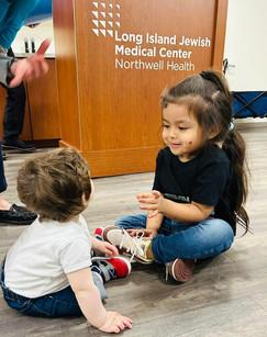 Two young children sit on a wooden floor, smiling and interacting. They are in front of a medical center sign. Casual and playful mood.