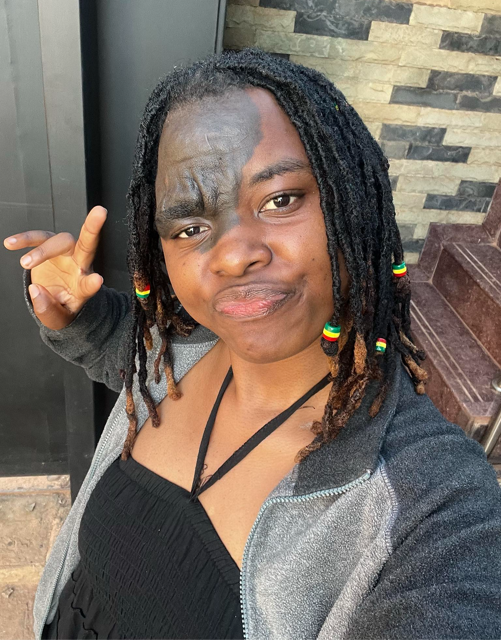 Person with dreadlocks and facial design makes a peace sign. Wears black top and gray jacket. Background has patterned stone wall. Smiling.