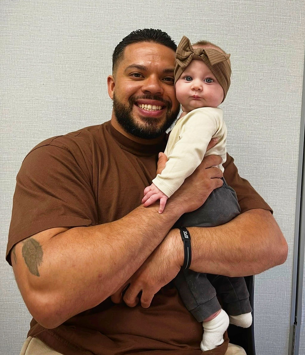 A father with brunette hair in a brown tshirt holds his baby girl lovingly. She has a large brown bow on her head and his smiley coyly at the camera. She has congenital melanocytic nevus on her scalp, gently tucked behind the brown bow.