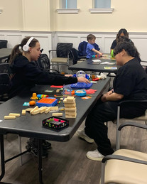 Two people play with toys at a table in a bright room, surrounded by colorful blocks and cards. A child in the background plays quietly.