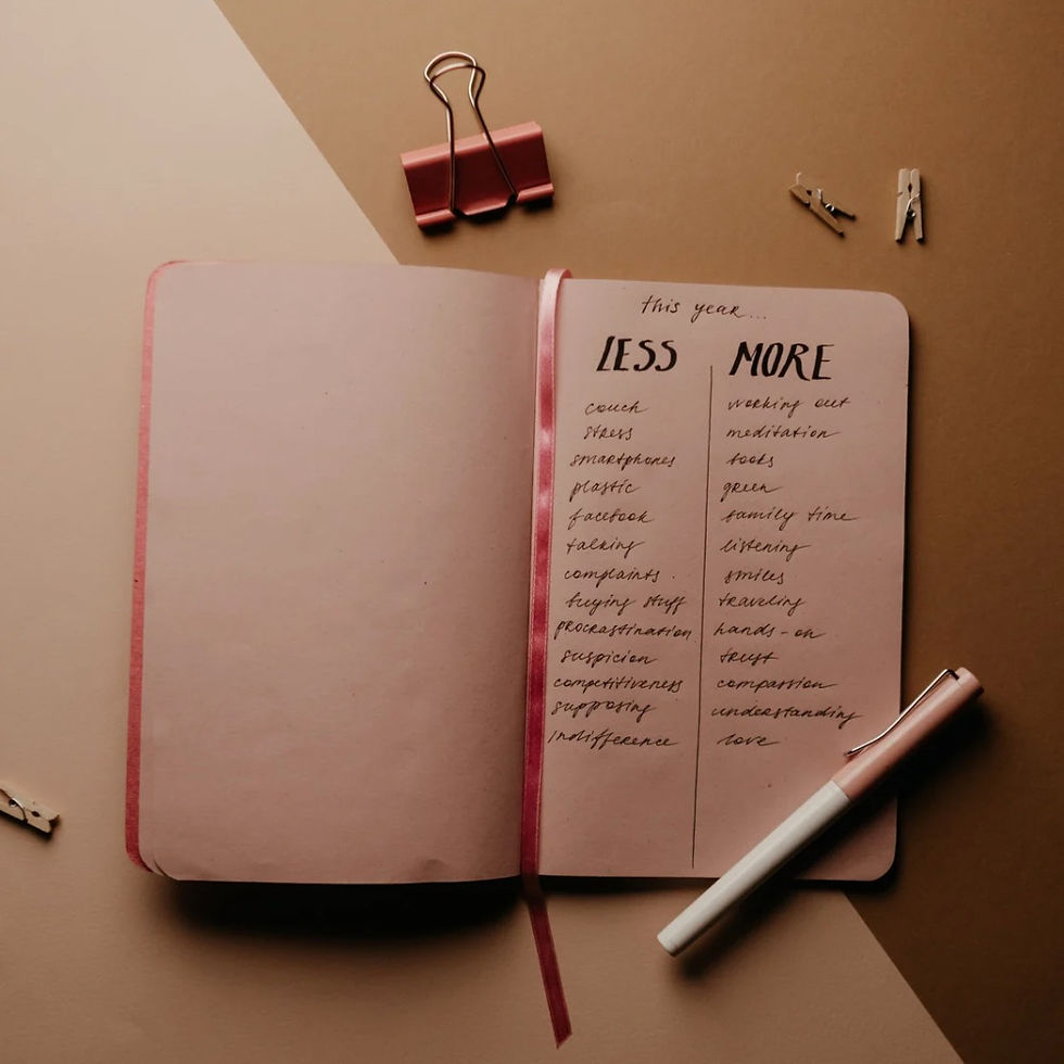 Open notebook with "Less" and "More" lists in cursive on pink pages. Pen and binder clips nearby on a beige surface. Relaxed mood.