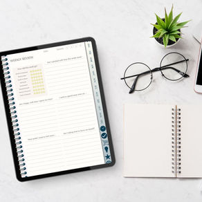Tablet showing a weekly review planner beside glasses, a blank notebook, and a small plant on a clean white desk.