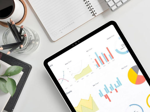 Tablet displaying colorful charts on a white desk with a keyboard, open notebook, jar of pencils, and coffee cup; organized workspace.