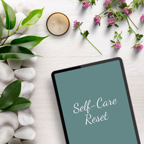 Tablet with "Self-Care Reset" text, surrounded by white stones, green leaves, purple flowers, and a lit candle on a light surface. Serene mood.