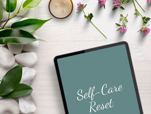 Tablet with "Self-Care Reset" text, surrounded by white stones, green leaves, purple flowers, and a lit candle on a light surface. Serene mood.