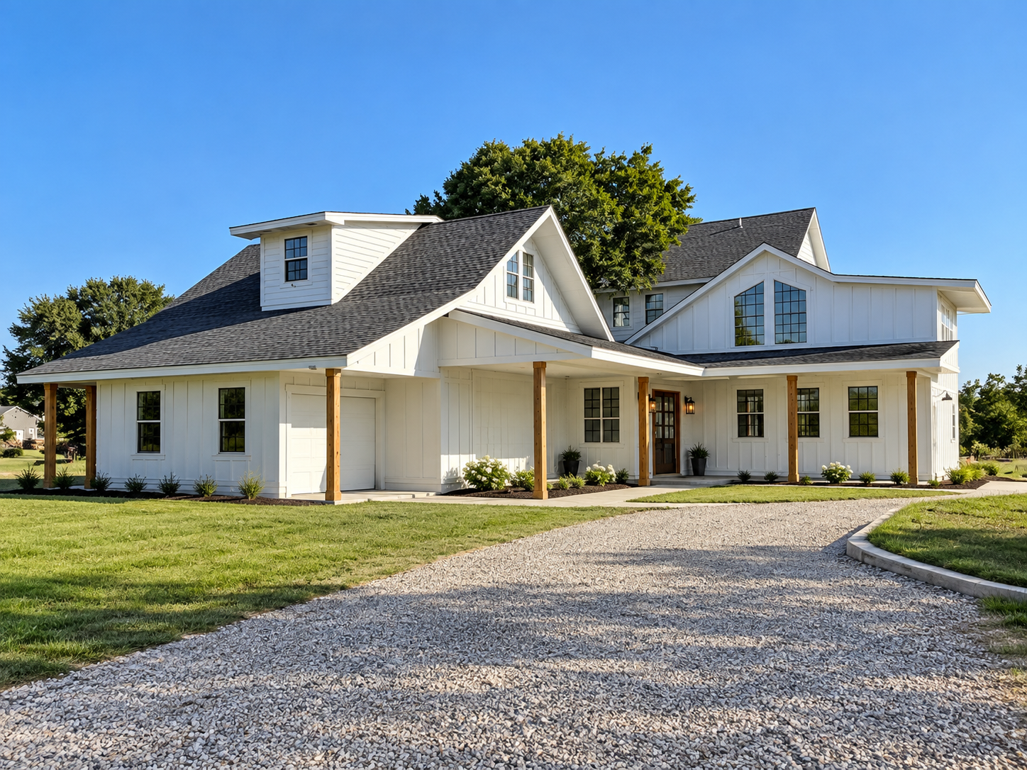 Wide landscape photo of a modern white farmhouse exterior on a sunny day, blue sky, green lawn, gravel driveway leading to the entrance, house centered in frame.