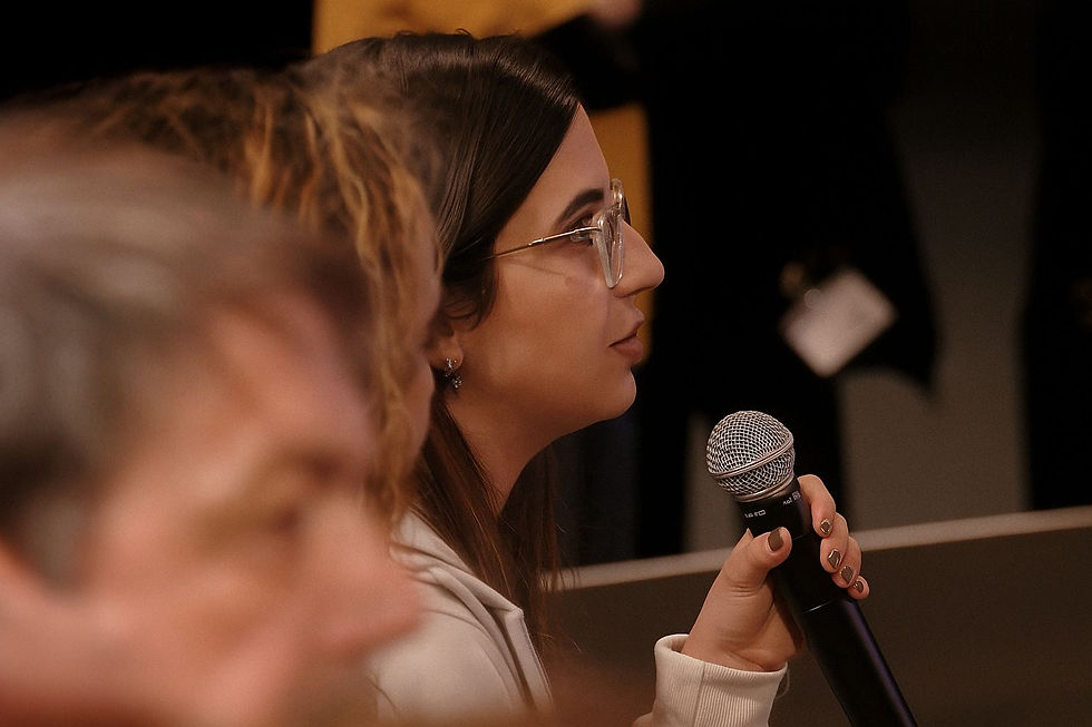 A woman in glasses holds a microphone, speaking attentively. Blurred man in foreground, dark background.