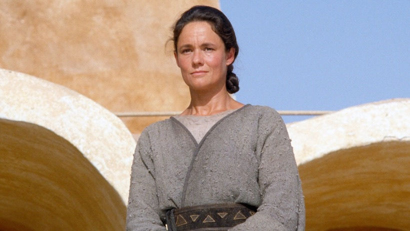 Woman in a gray robe stands confidently outside against a tan stucco building under a clear blue sky. She appears calm and composed.