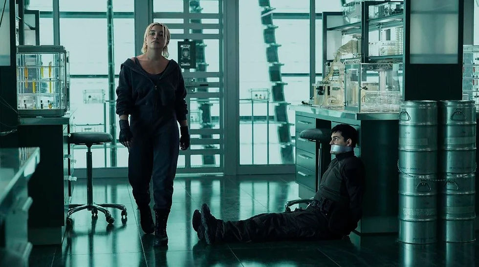 Woman in black clothing walks in a lab. A man is sitting, tied and gagged. Stainless steel tanks and lab equipment in the background. Mood is tense.