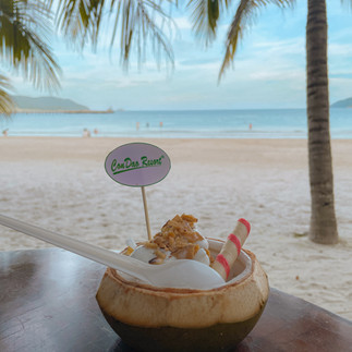 A coconut cream on a table with a sandy beach and calm sea in the background in Con Dao.