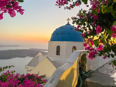 Blue-domed churches and whitewashed buildings in Santorini at sunset, with bougainvillea and cliffside views over the Aegean Sea