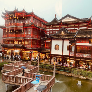 Visitors holding umbrellas walk across zigzag bridges at Yuyuan Old Street, surrounded by brightly lit traditional Chinese buildings on a rainy evening.