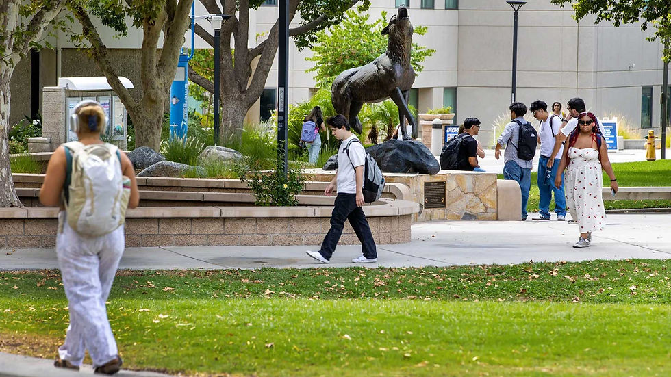 Estudiantes caminan por un campus junto a una estatua de caballo; césped, árboles y edificio claro al fondo.