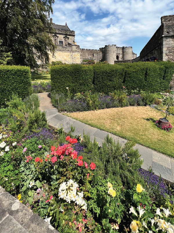 Stone castle ruins in the background with a garden pathway bordered by colorful flower beds and trimmed hedges.