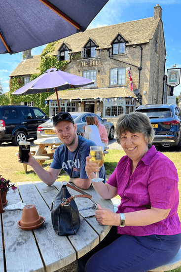 A man and woman sitting at an outdoor pub table, raising glasses of beer and cider, with The Bell Inn’s stone building in the background.