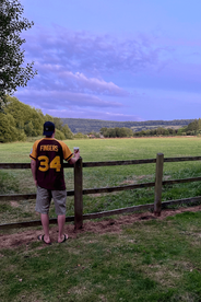 A man wearing a sports jersey leans against a wooden fence, gazing at rolling hills under a purple-tinted sky.