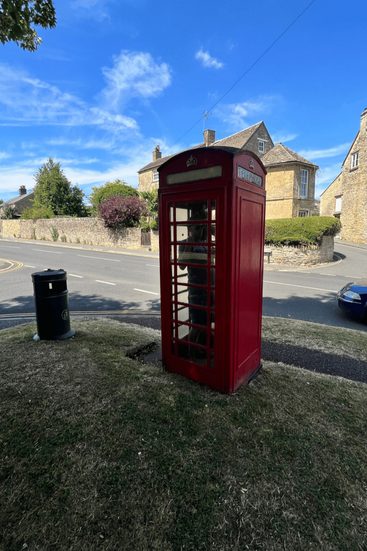 A traditional red telephone booth on a grassy roadside, with stone houses in the background under a bright blue sky.