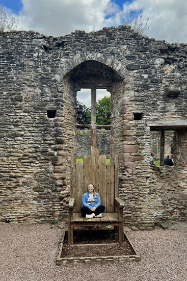 A woman sitting on a large wooden throne within the ruins of Ludlow Castle, framed by high stone walls.