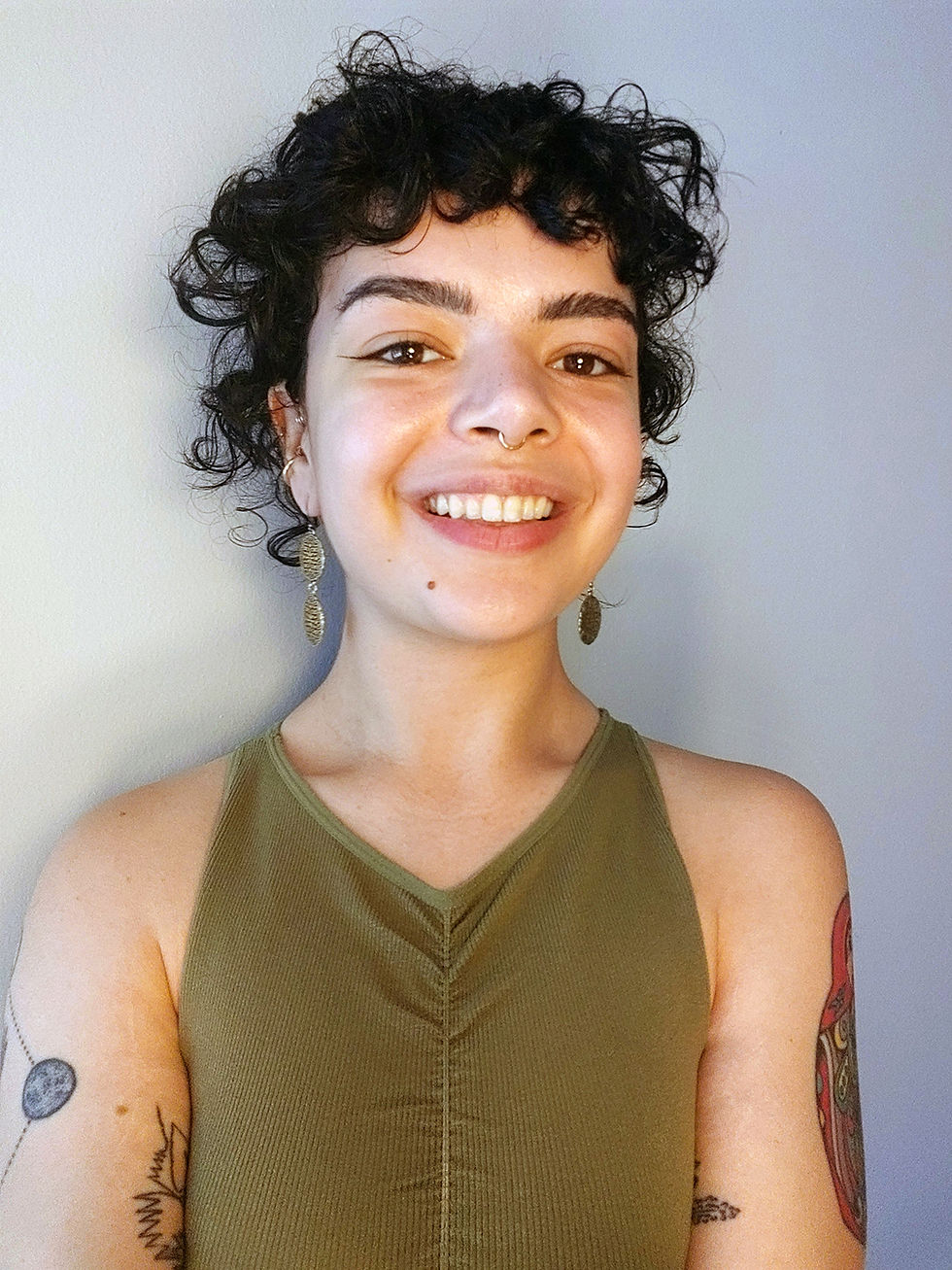 A headshot style photo of Lexi Falek, One Yoga's new Membership and Engagement Director. Lexi has short dark curly hair, is wearing earrings and a green tank top, and has tattoes on her bare arms.