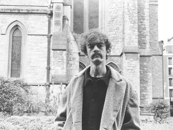A young man with messy hair and a solid mustache, indie vibes
