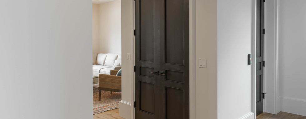 Hallway with wooden floor leads to dark double doors. Through an open door, a beige sofa is visible in a softly lit room. Calm ambiance.