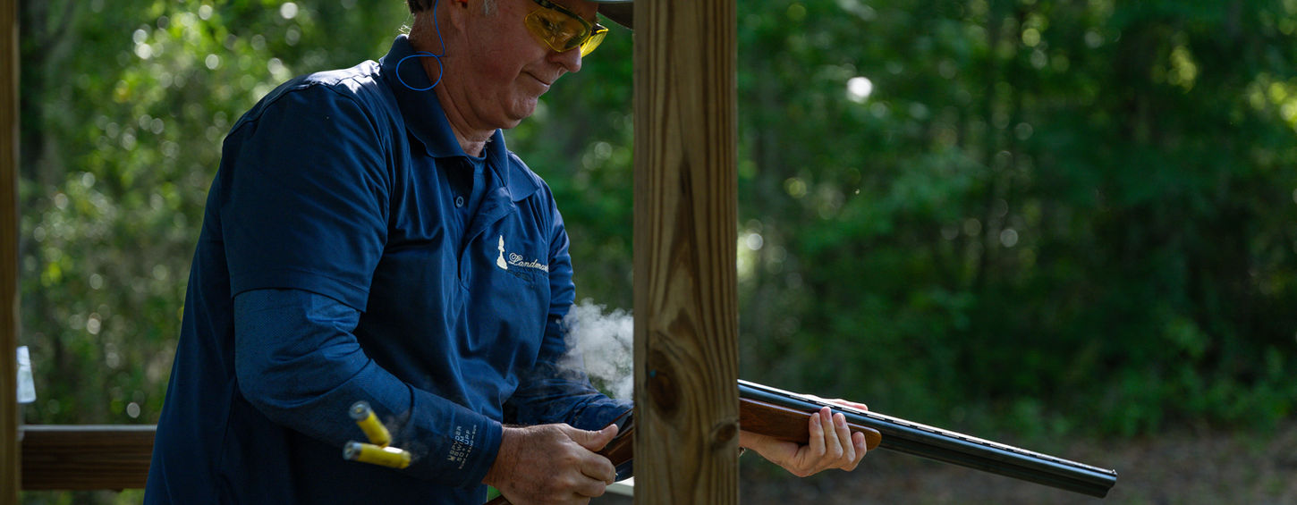 A man wearing a cap, safety glasses, and ear protection opens a shotgun at a wooden shooting station after firing. Two spent shells eject from the gun with a small puff of smoke, and a box of ammunition sits on the ledge, with trees in the background.
