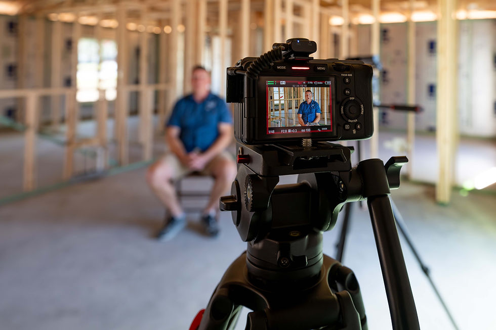 Cámara en trípode grabando a Brain Smallwood, propietario de Belac Homes, sentado en un edificio en construcción. El monitor muestra al sujeto con camisa azul. Luz natural brillante.