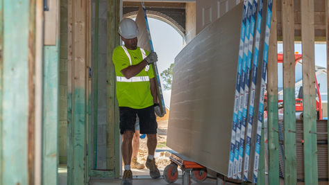 Construction worker carrying a large sheet of drywall into a framed residential home under construction with stacked drywall panels on a cart nearby.