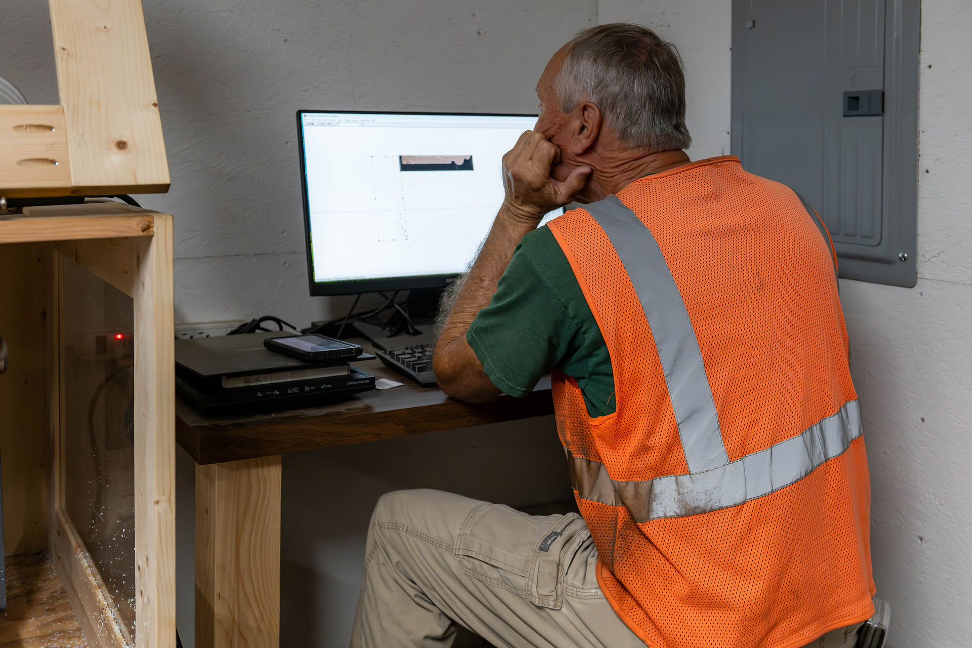 Man in orange safety vest works at desk with computer, focused on screen displaying design. Wooden furniture and equipment visible.