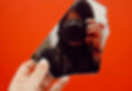 A hand holds a broken mirror piece reflecting a person with a camera. Bright red background enhances the image's intensity.