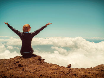 Person sitting on rocky peak, arms raised in joy, overlooking clouds against a bright blue sky, conveying a sense of freedom and triumph.