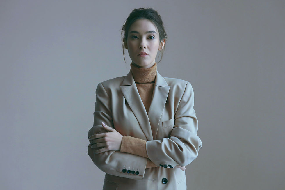 Woman in a beige blazer and tan turtleneck, crossing arms, stands against a plain background, exuding a calm and confident mood.