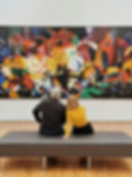 Two people sit on a bench, admiring a vibrant abstract painting with geometric patterns and bold colors in a gallery setting.