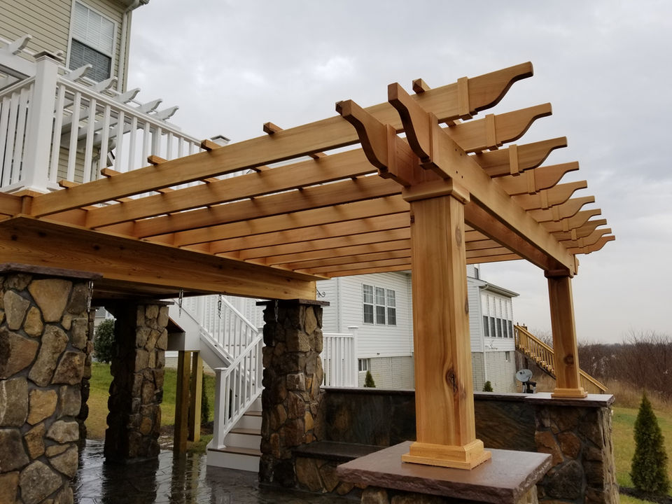 Wide view of a composite deck with vinyl railings leading down to a patio with a cedar pergola and real stone columns.