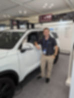 CEO Tim Balz is standing in the  Hyundai Motor Group Open Innovation Lounge next  and leaning on a white Hyundai SUV wearing his Kalogon polo and khaki pants.