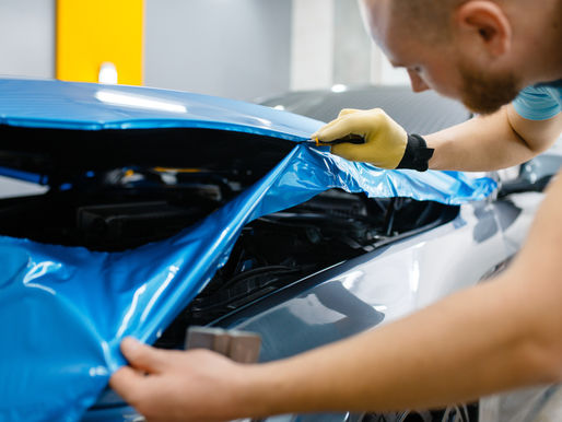 Man applies blue car wrap in garage, using tool and wearing gloves. Close-up of focused work with reflective surface. Yellow post visible.