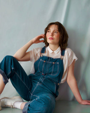 Jessy in blue overalls leaning on her knee with a white collard shirt underneath. She is looking into the distance.