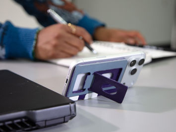 CARD by  LUVO. centered on the table and being used as a phone stand with a man sketching in the background and the left back corner of a laptop in the left foreground.