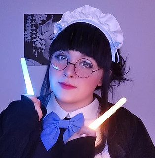 Maid Kumo is posing with two light sticks beside her face. The lights shinning on her face. 