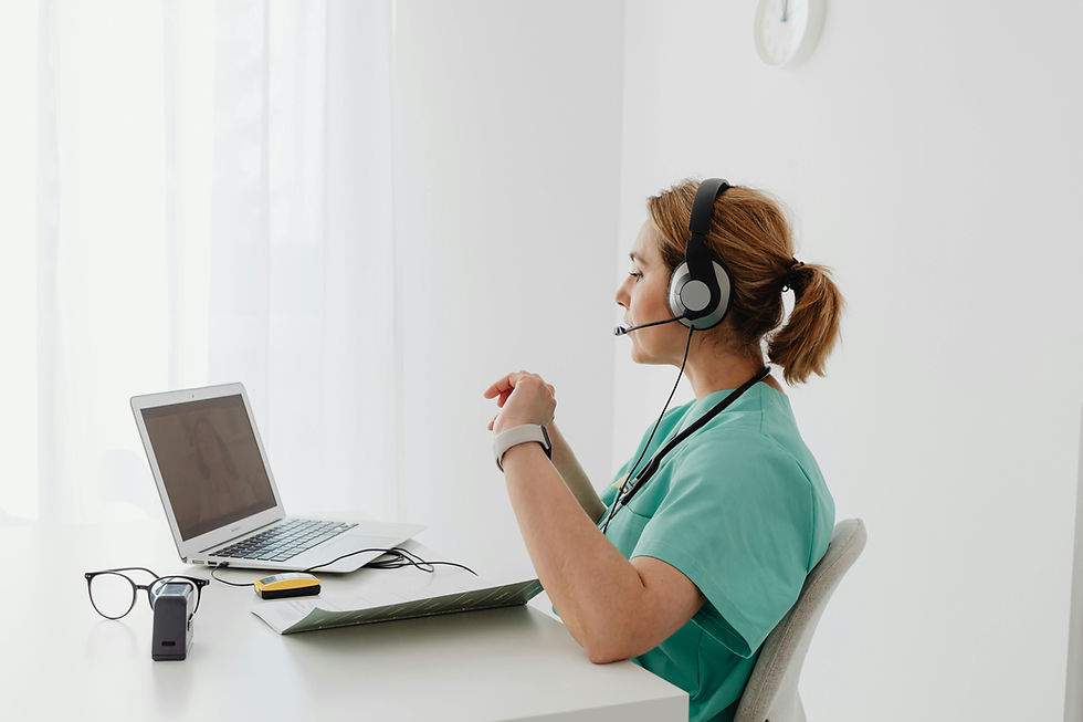 Nurse in green scrubs with headset sits at a desk, video chatting on a laptop in a bright, white room. Glasses and papers lie nearby.