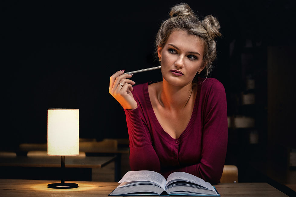Woman in maroon sweater pensively holds a pen, near an open book in dim lamp-lit setting. She appears thoughtful.