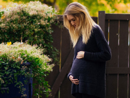Pregnant woman in a black sweater holds her belly while standing by a wooden fence. Lush green bushes and autumn leaves surround her.