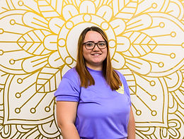 Yoga instructor, holly, with long dark hair and dark glasses, wearing a puple branded tshirt stood infront of a golden mandala.